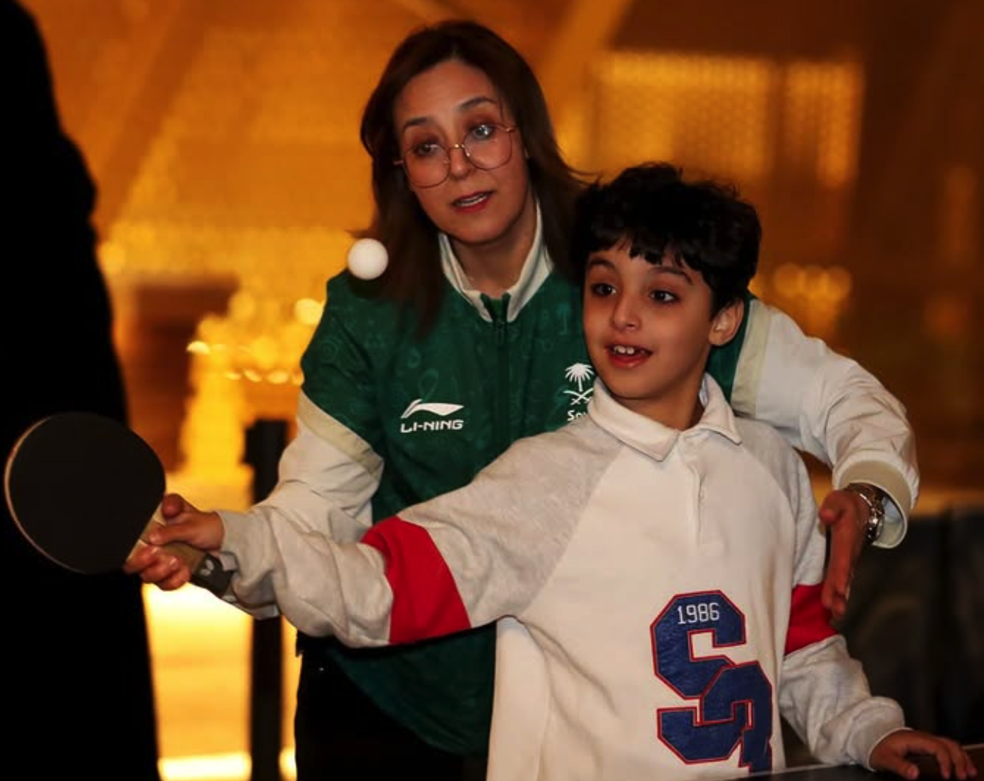 Saudi table tennis players - woman in national team jersey with young player, representing growth and development in Saudi Arabia